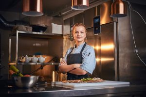 Confident and serious female chef standing with hands crossed in a dark kitchen next to cutting board with vegetables on it