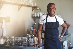 Handsome black entrepreneur stands by cafe counter lined with jars of tea while wearing dark colored apron