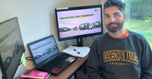 Man seated in front of computer, laptop and pink calculator.