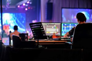A person is sitting in front of multiple monitors. A stage is lit up in front of them for an event.