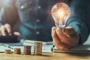 There is a stack of coins on a desk with a lightbulb next to it. A man is cropped in this image holding the lightbulb.