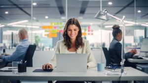 A woman is sitting and working on a laptop in an office.