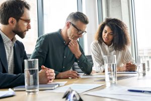 Three professionals engaged in a serious discussion around a conference table with documents and water glasses.