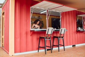 A red pop-up restaurant has an open window with chairs for people to sit and eat.