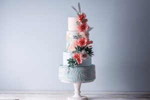 A four-tiered wedding cake with pink flowers is resting on a cake stand with a white background.