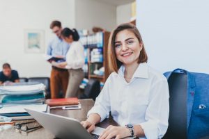A smiling professional works on a laptop at her desk, with colleagues discussing documents in the background.