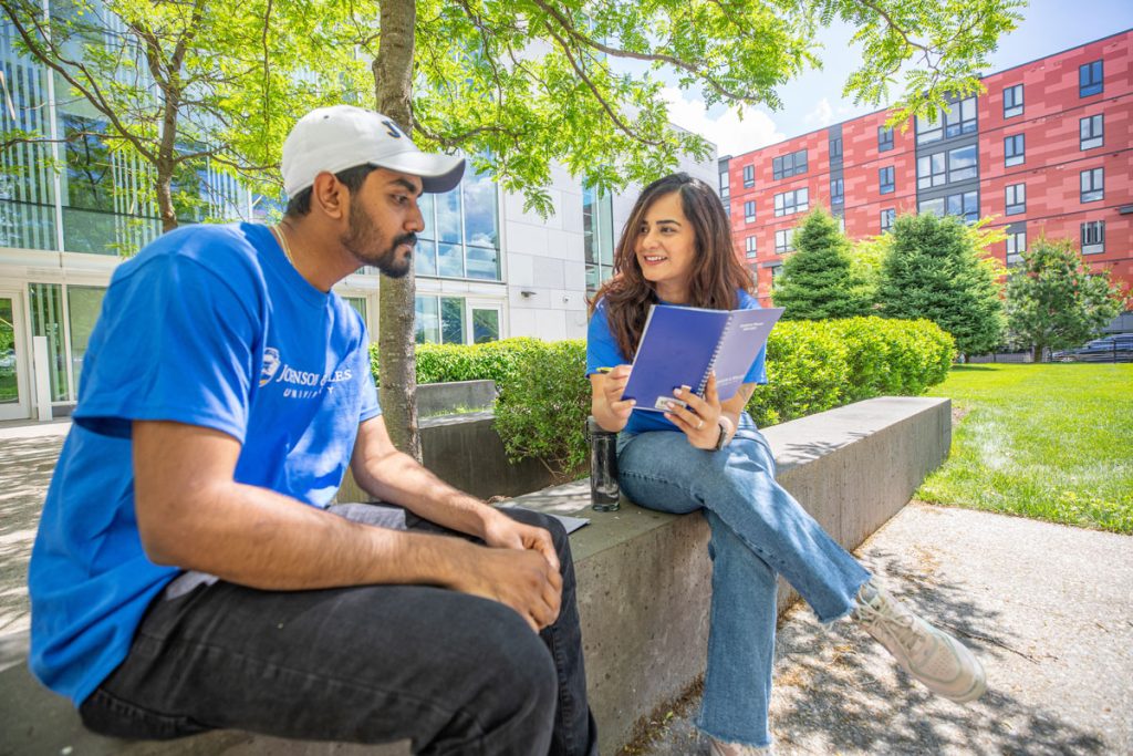 A male student in a white cap and a female student, both wearing blue T-shorts with JWU logos, are seating on a wall while talking together in an outdoor campus courtyard.
