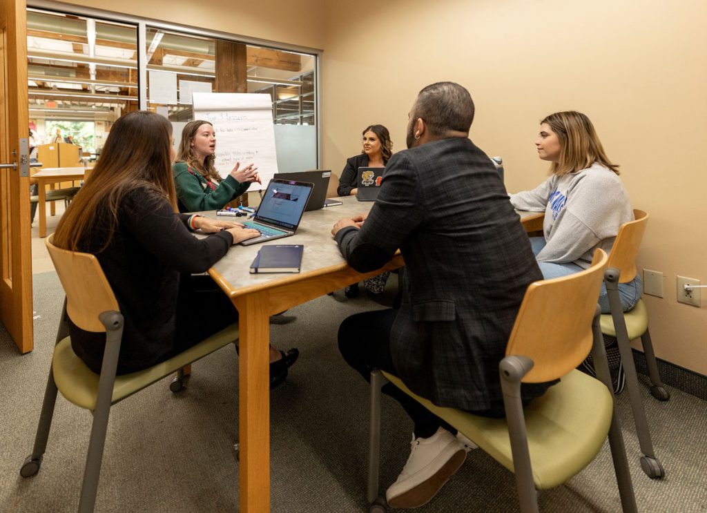 One male and three female students with laptops are seated at a table in a campus library, listening to a woman with a laptop and presentation pad speaking to them.
