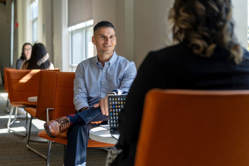 A man wearing a light blue patterned button-down shirt, blue pants and brown shoes is sitting across from a woman in a campus lounge area.
