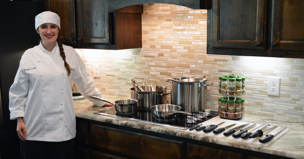 JWU online culinary student in uniform standing in kitchen with culinary tools and equipment.