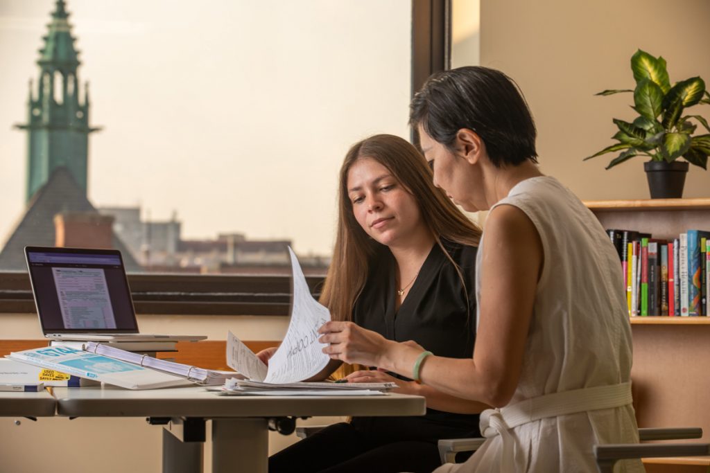 A university student and staff professional, seated at a table in an office which has a view out to other campus buildings, review financial forms together.