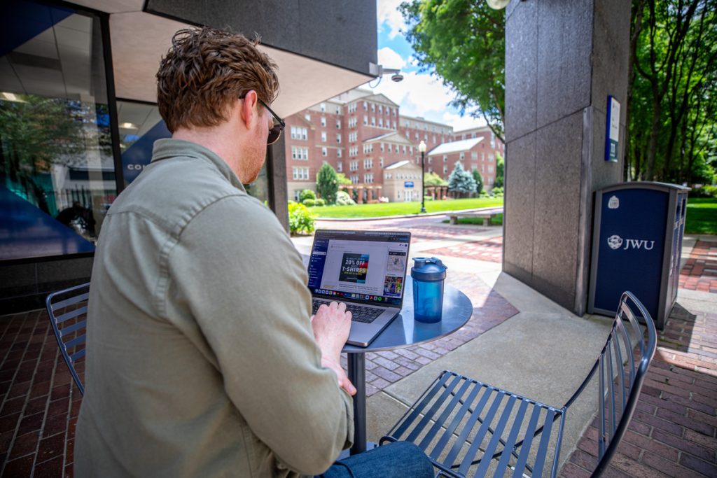 View over the shoulder of a male adult student who is seated at a table on an outdoor campus patio, looking at a laptop screen.