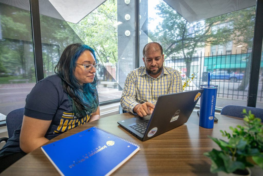 Young woman and young man seated at table in front of windows with views out to the city street, looking at the young man's laptop screen.