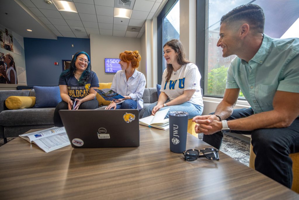 Three young women and one male, seated around a table with open books and a laptop smiling and talking with one another, suggesting a study session.