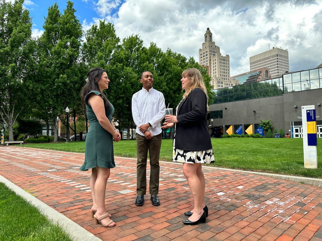 Professionally dressed group of two young women and one young man conversing while standing on a brick walkway in a campus quad.