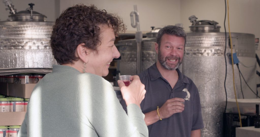 Customer holding a small sample cup of cider while a smiling owner, Jay Wiedenheft. stands in front of cider making equipment.