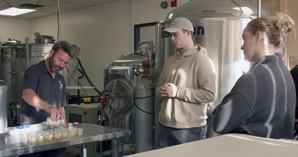 Backed by cider making equipment, Jay Wiedenheft pours small sample cups from a cider can, while two JWU students watch.