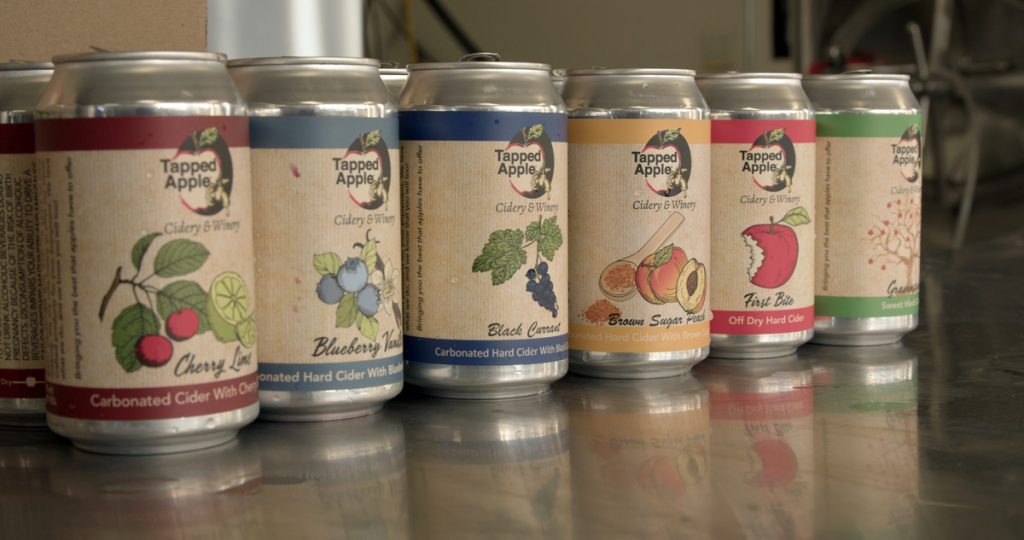 A display of 6 cider cans, each labeled with a colored illustration of the name of and the fruit in each cider blend.