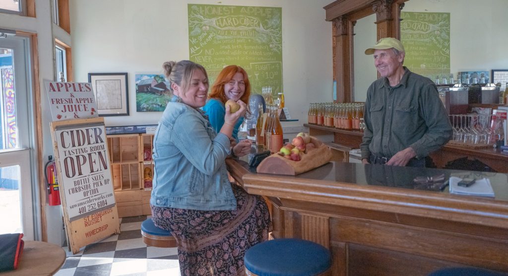 Two women seated at a cider bar examine an apple, while a gentleman in a yellow cap and work shirt stands behind the bar counter and looks on.