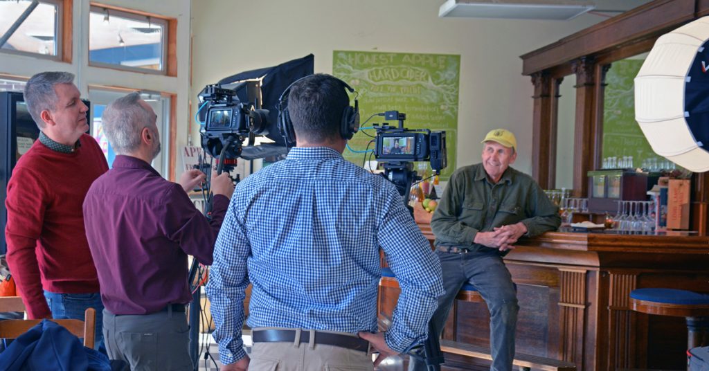 Three men stand behind a professional video camera focused on a gentleman in a yellow cap, work shirt and jeans, seated on a barstool in front of his cider bar.