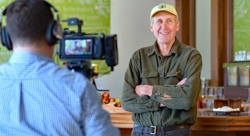 Back of the head of cameraman as he peers through the viewfinder of his video camera at Spence Morris, in yellow cap and work shirt, smiling and with hands across his chest.