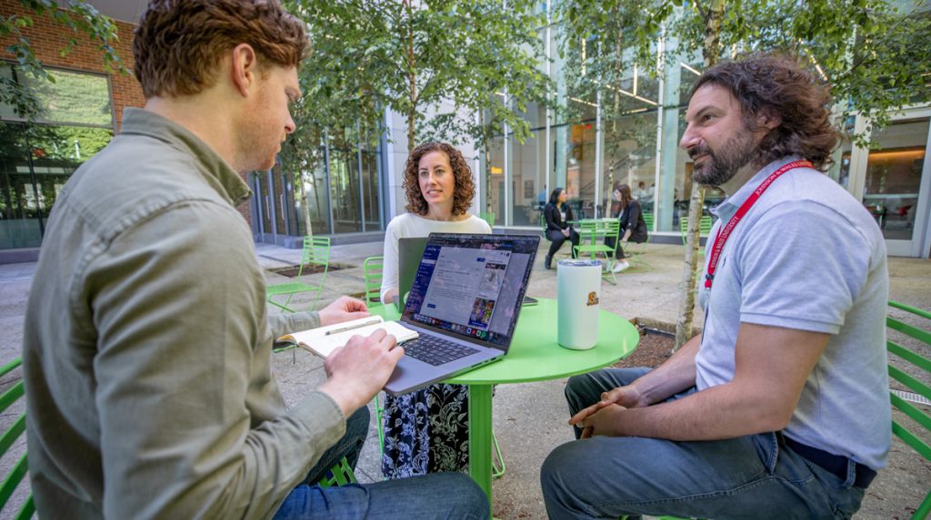 A male adult student with a laptop and another adult male student converse while seated at a table in an outdoor area, and a female adult student seated across from them listens.