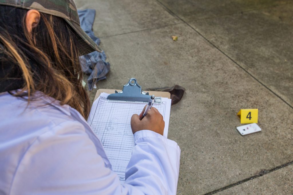 Viewing over the shoulder of a young woman in a white lab coat and holding a clipboard with a chart in which she is entering data about a piece of evidence on the sidewalk of a crime scene.