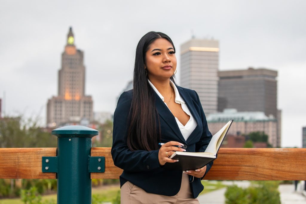 Young woman writing in a notebook, while standing outdoors with a view of city buildings in the background.