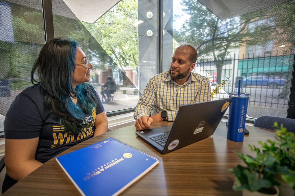 Two adult students of mixed gender seated in a lounge area with a notebook and laptop on the table.