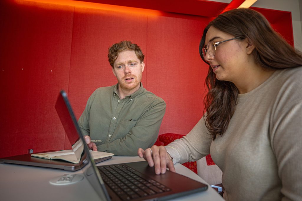 Female adult student working on her laptop in the foreground shares her screen with a male adult student.