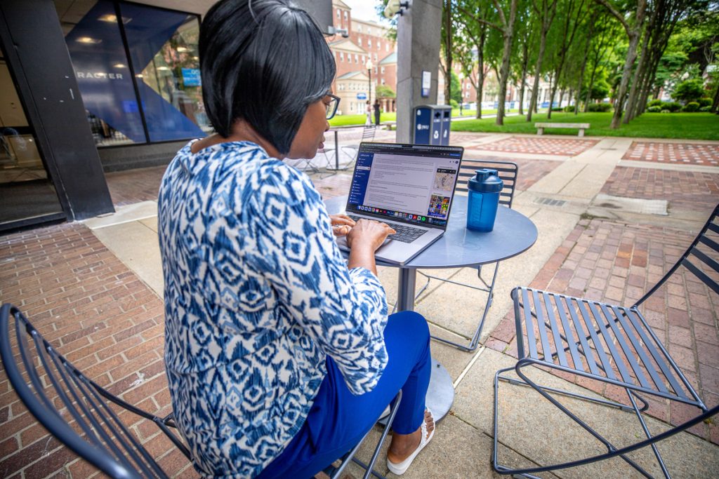 Over the shoulder view of a female adult student seated at a cafe table in a courtyard, working on her laptop.