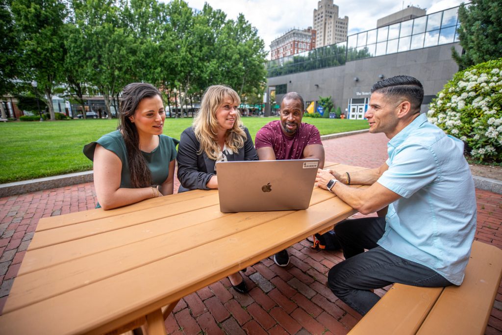 Four adult students of mixed genders and ethnicities seated at a picnic table outdoors, viewing a laptop screen.