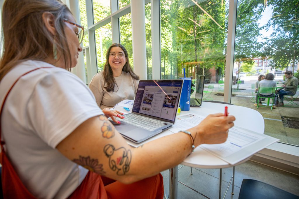 A female adult student with a laptop and a second female adult student with a book, seated at a table in front of windows looking out to a courtyard.