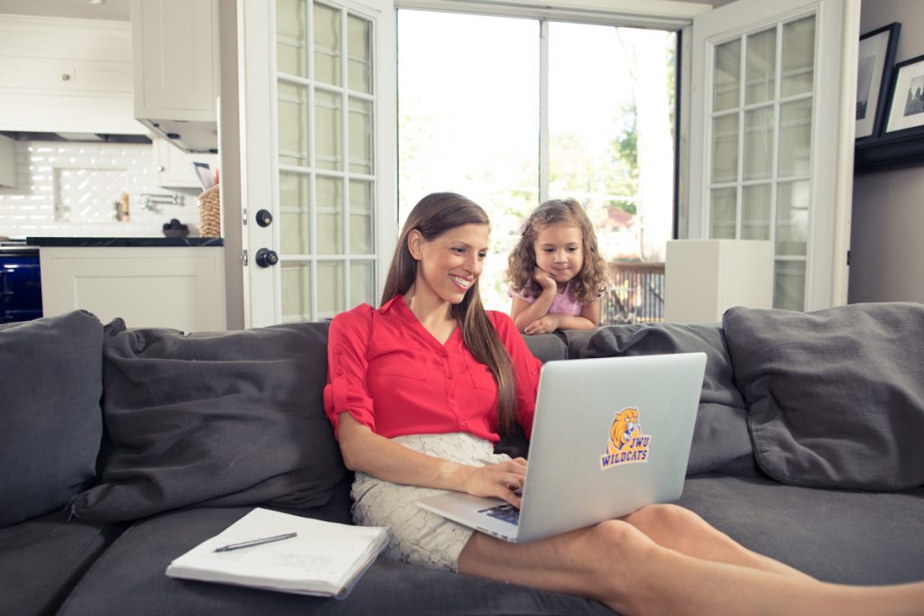 Adult female student working on laptop while seated on couch at home and young daughter looks over her shoulder at the the laptop screen.