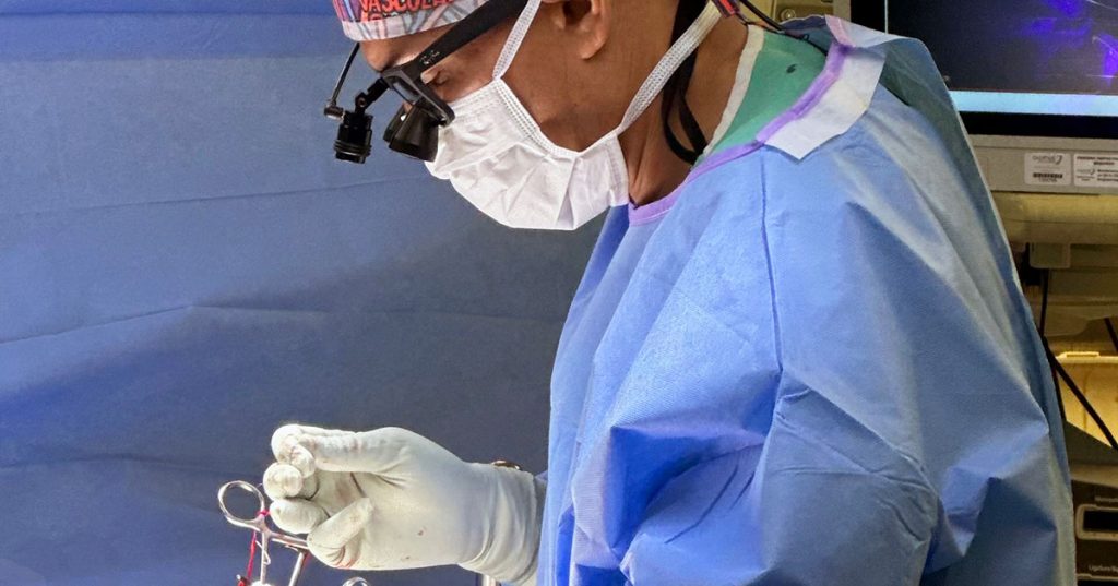 Dr. Ajay Patel in a surgical mask, gown, wearing a surgical loupe and holding scissors with gloved hands during an operation.