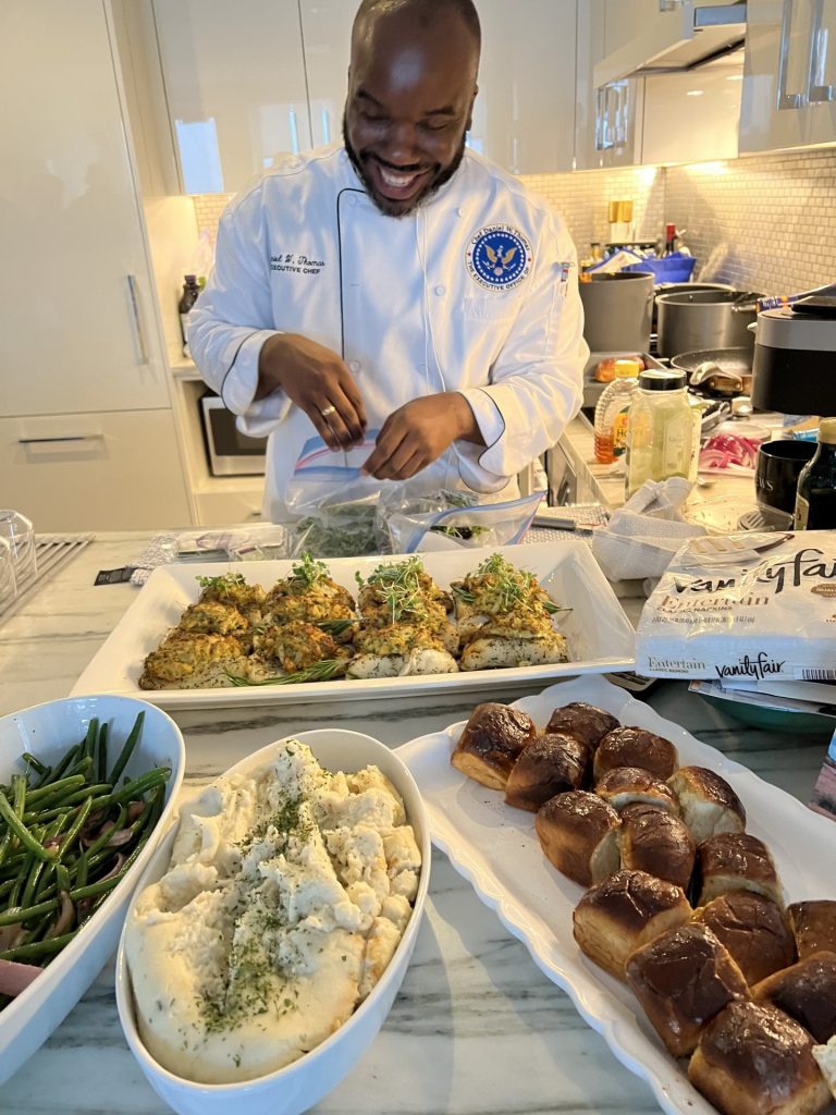 A smiling Daniel Thomas in his chef's uniform admiring the spread of food he has prepared.