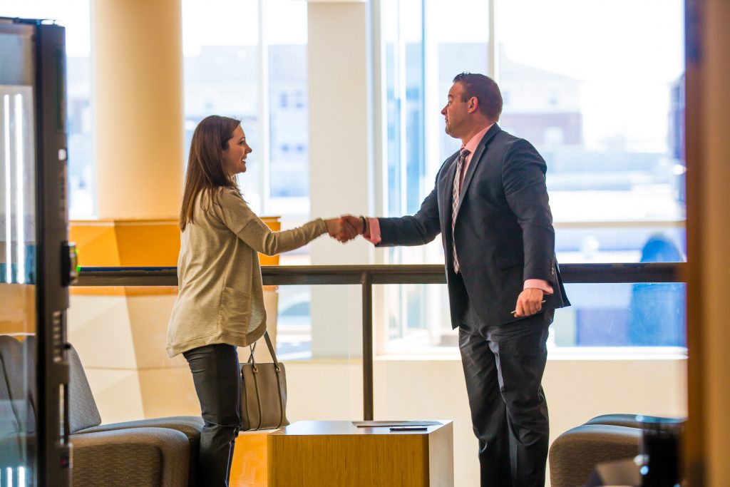 A young woman shakes the hand of a man in a business suit as they stand in front of windows overlooking the city of Providence, RI.