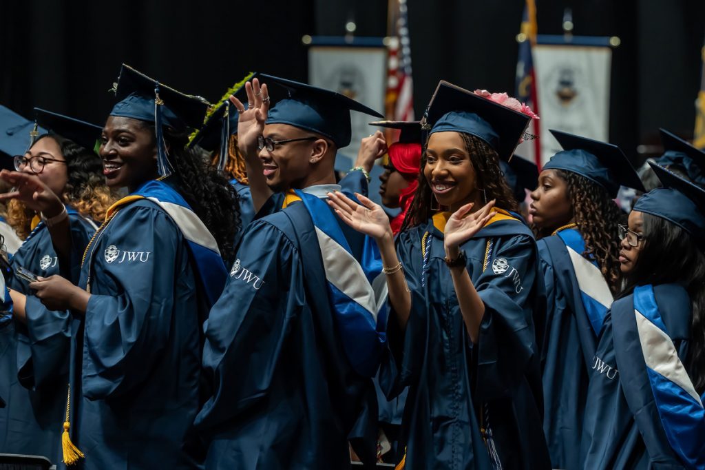 Five women an a man of color wearing their caps and gowns wave as the walk in the procession at the Johnson & Wales 2025 Commencement in Charlotte, NC.