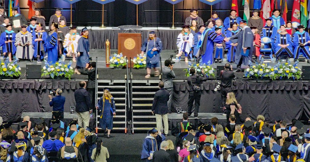 Faculty and leadership seated on the stage during the undergraduate ceremony, while students walk across to receive their diplomas.