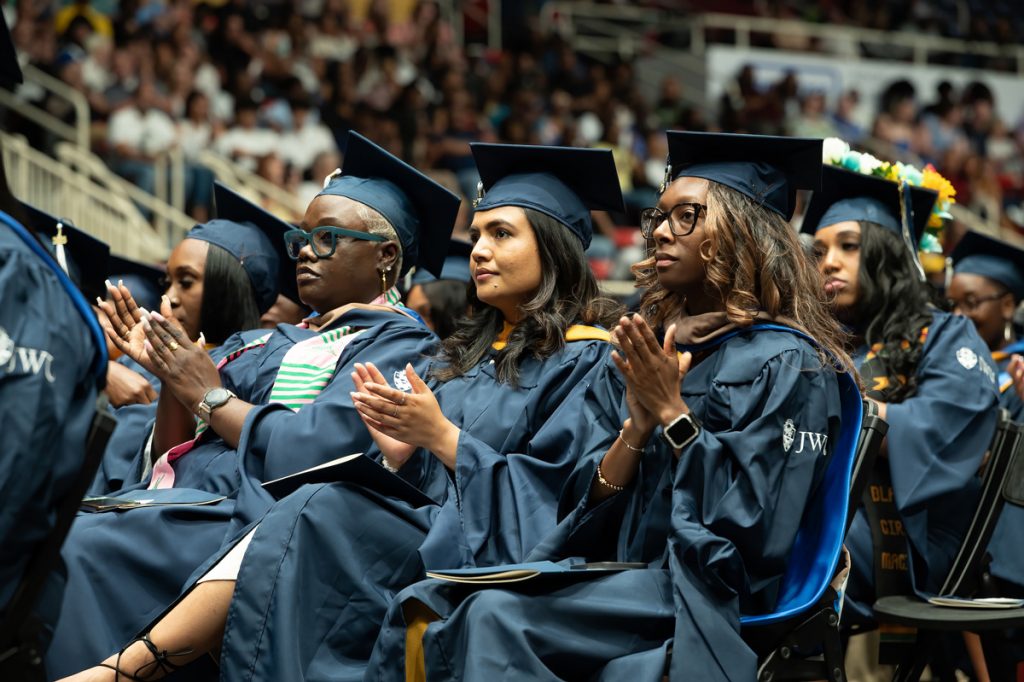 Five women of color wearing their caps and gowns applaud a speaker at the Johnson & Wales 2025 Commencement in Charlotte, NC.