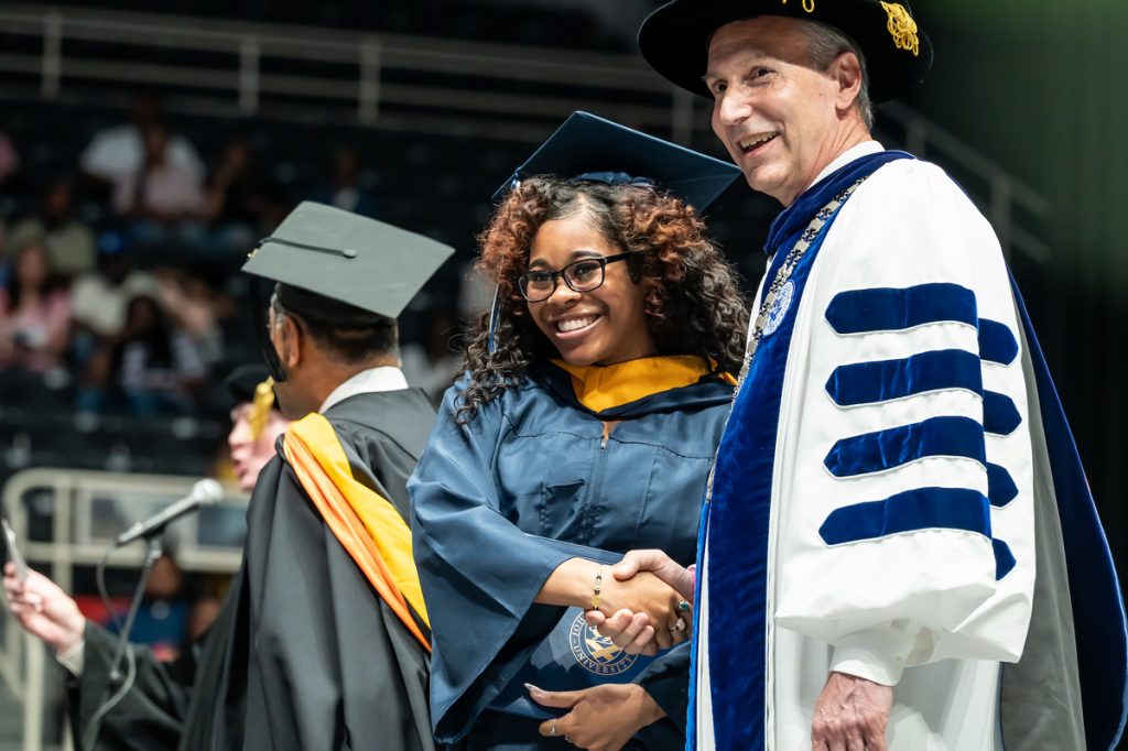 President of the JWU Charlotte campus, a mature man in his robes, shakes the hand of a graduate who has just accepted her diploma at the 2025 Johnson & Wales Commencement in Charlotte.