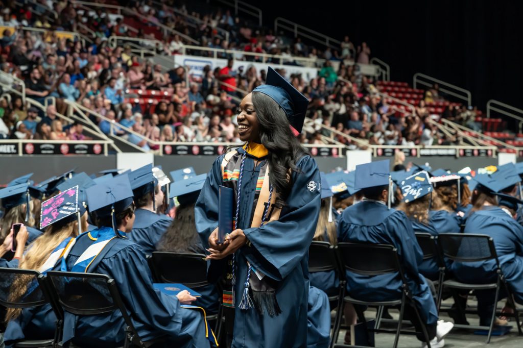 Woman of color in a cap and gown walks past rows of graduating students at the Johnson & Wales 2025 Commencement in Charlotte.