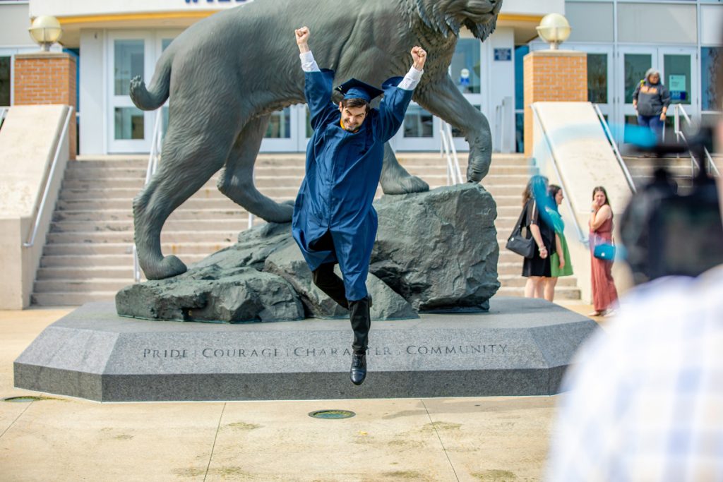 Smiling male graduate in a cap and gown jumps for joy with arms raised over his head in front of the Wildcat statue on the Harborside campus in Providence during the 2025 Senior Toast.