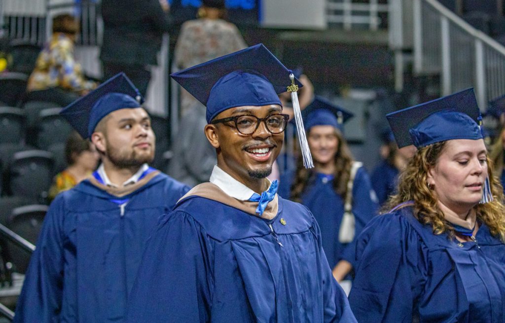 Graduating students of mixed gender and background, in their caps and gowns, with one male smiling at the camera at the 2025 Johnson & Wales Commencement in Providence.
