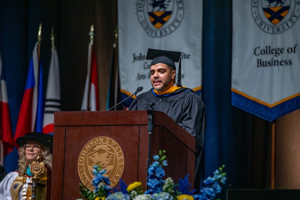 Male speaker in a cap and gown at the podium during the 2025 Johnson & Wales Commencement in Providence.