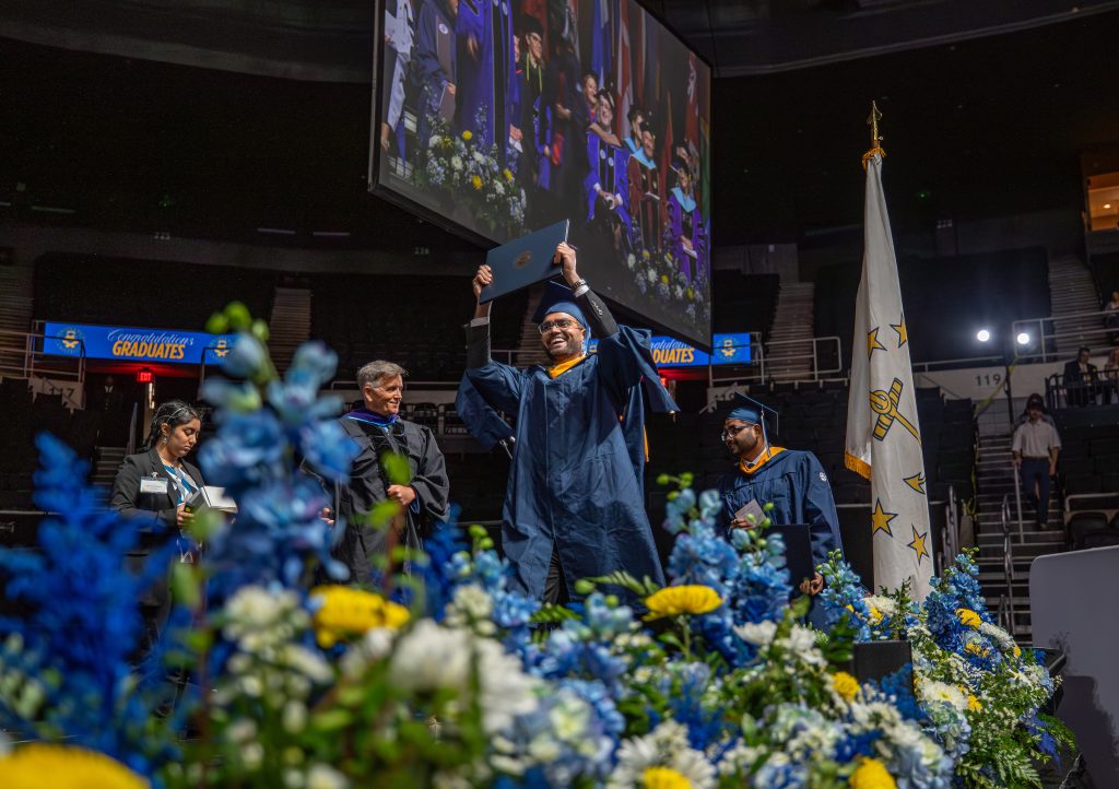 Shot of male student holding his diploma above his head and smiling with floral arrangements in the foreground at the 2025 Johnson & Wales Commencement in Providence.