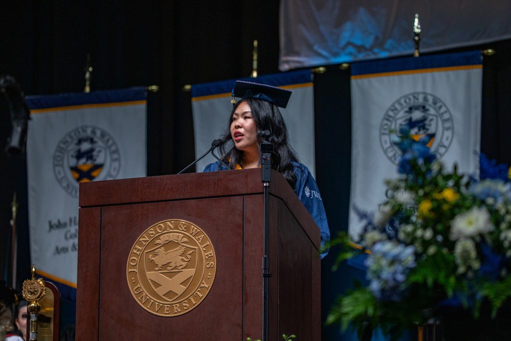 Young woman of Asian descent in cap and gown giving a speech at the podium at the 2025 Johnson & Wales Commencement in Providence for undergraduate students.