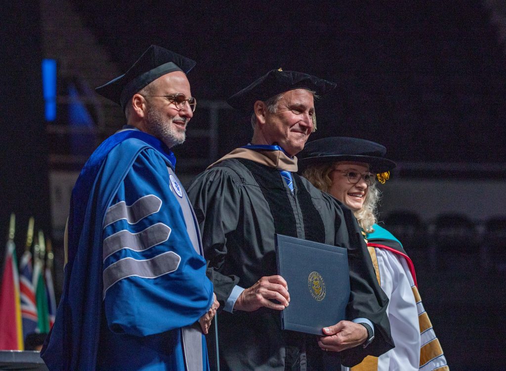 Adult male in robes holding an honorary degree, flanked by Johnson & Wales Provost on the left and Chancellor on the right at the 2025 Undergraduate Commencement in Providence.