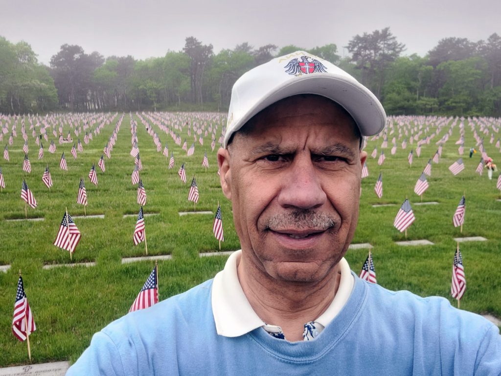 U.S. Navy veteran in his fifties, wearing a cap, standing in a military cemetery that has rows of graves marked with American flags.
