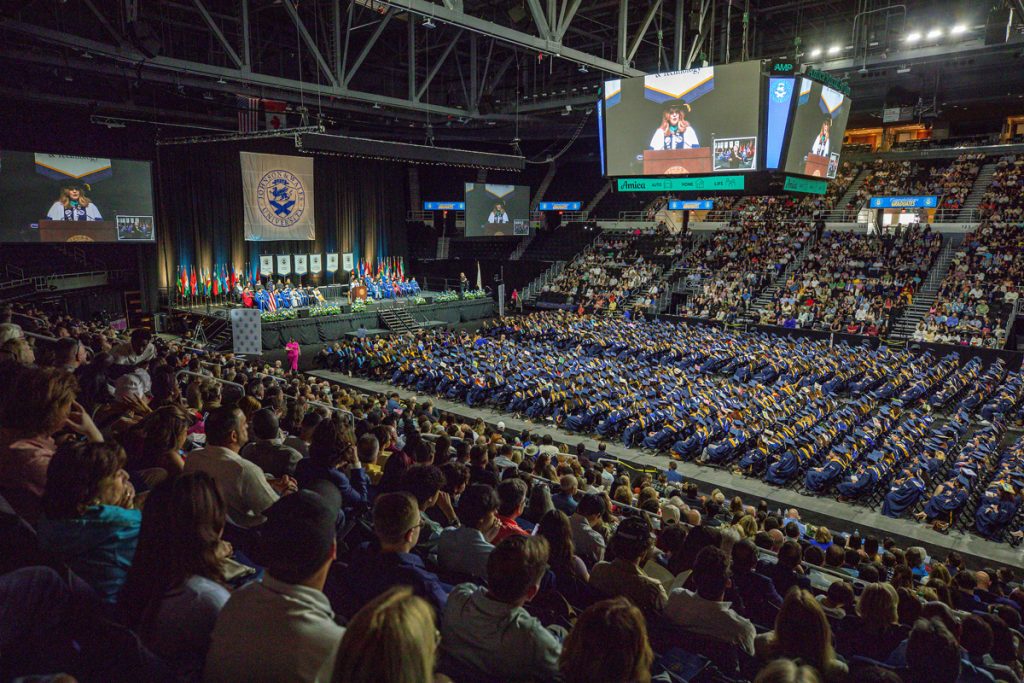 Shot overlooking seated guests and rows of graduates to the screen televising the graduation ceremony at the 2025 Johnson & Wales Undergraduate Commencement ceremony in Providence.